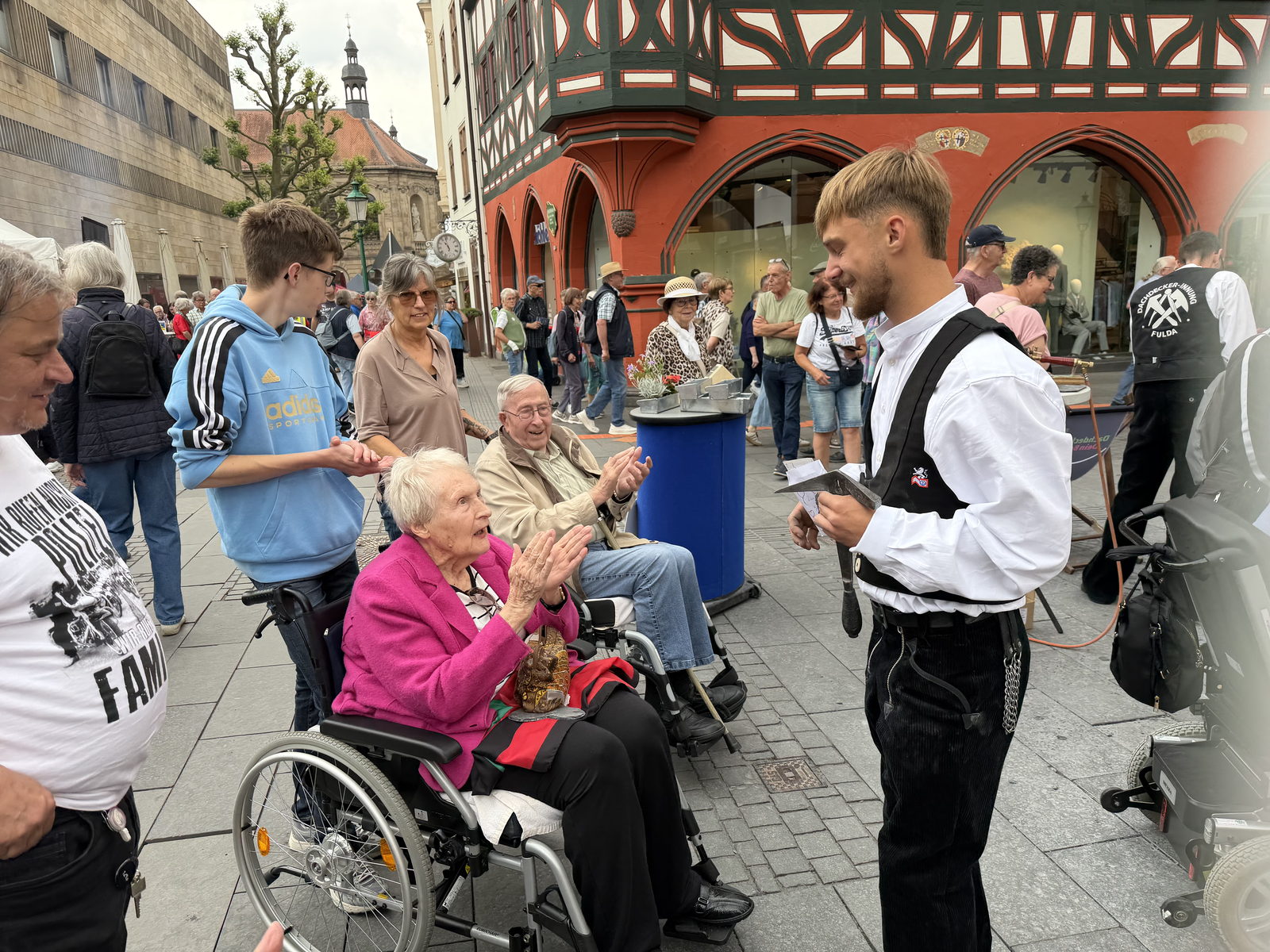 Bewohnerinnen und Bewohner im Rollstuhl applaudieren einem jungen Dachdecker in Zunftkleidung auf dem Fuldaer Marktplatz