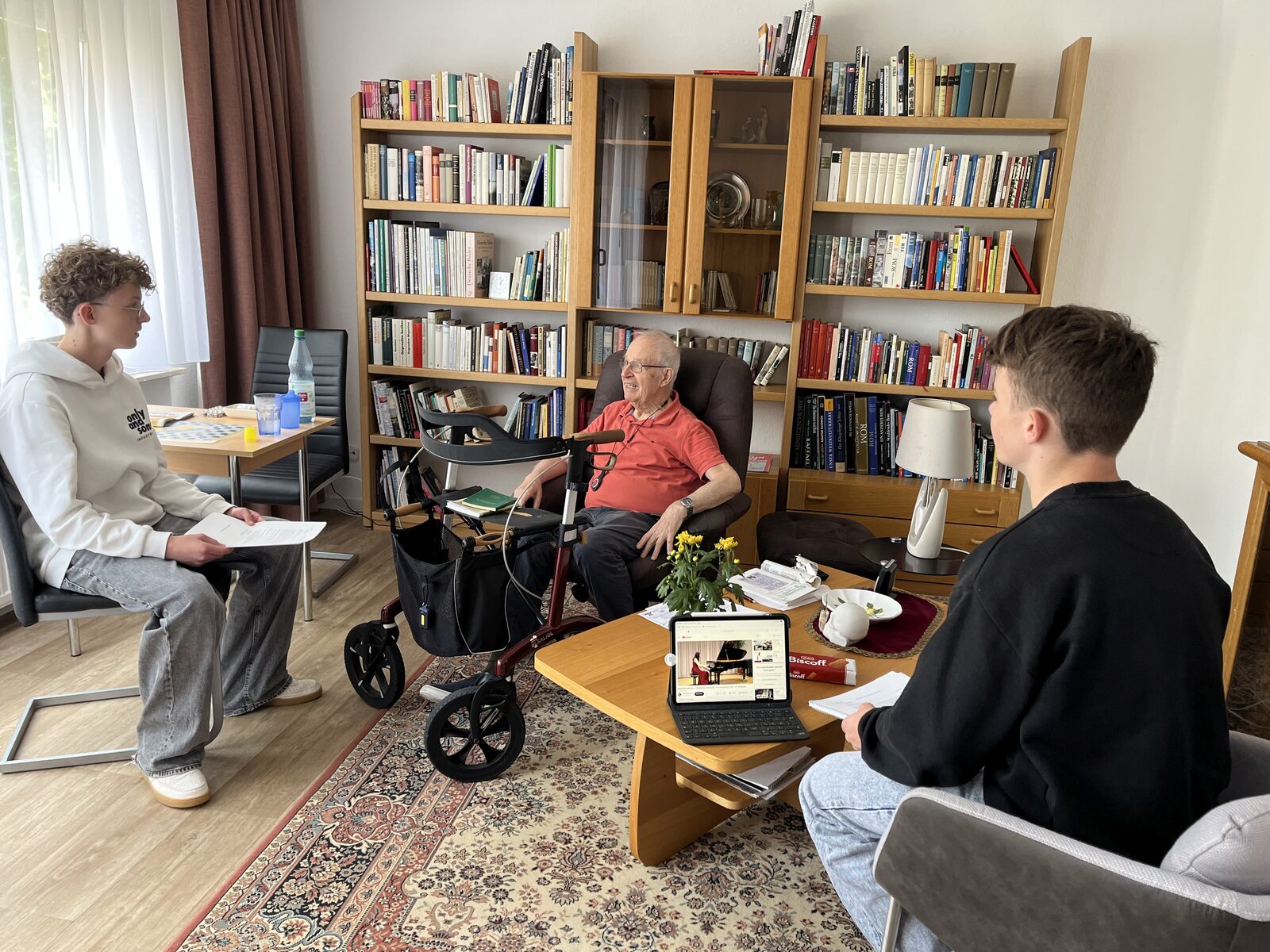 Zwei Schüler befragen einen Bewohner des Mediana Wohnstifts in seinem Apartment vor einem Bücherregal zu seinen Musikvorlieben
