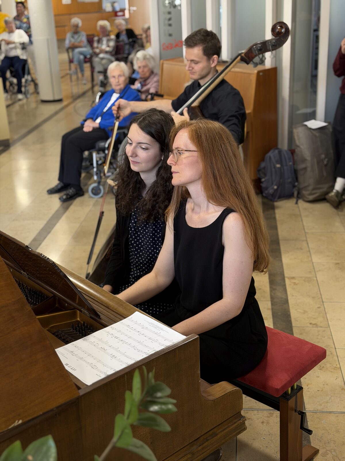 Zwei Pianistinnen spielen vierhändig Klavier, dahinter ein Kontrabassist, im Foyer des Mediana Wohnstifts hören Bewohnerinnen und Bewohner zu